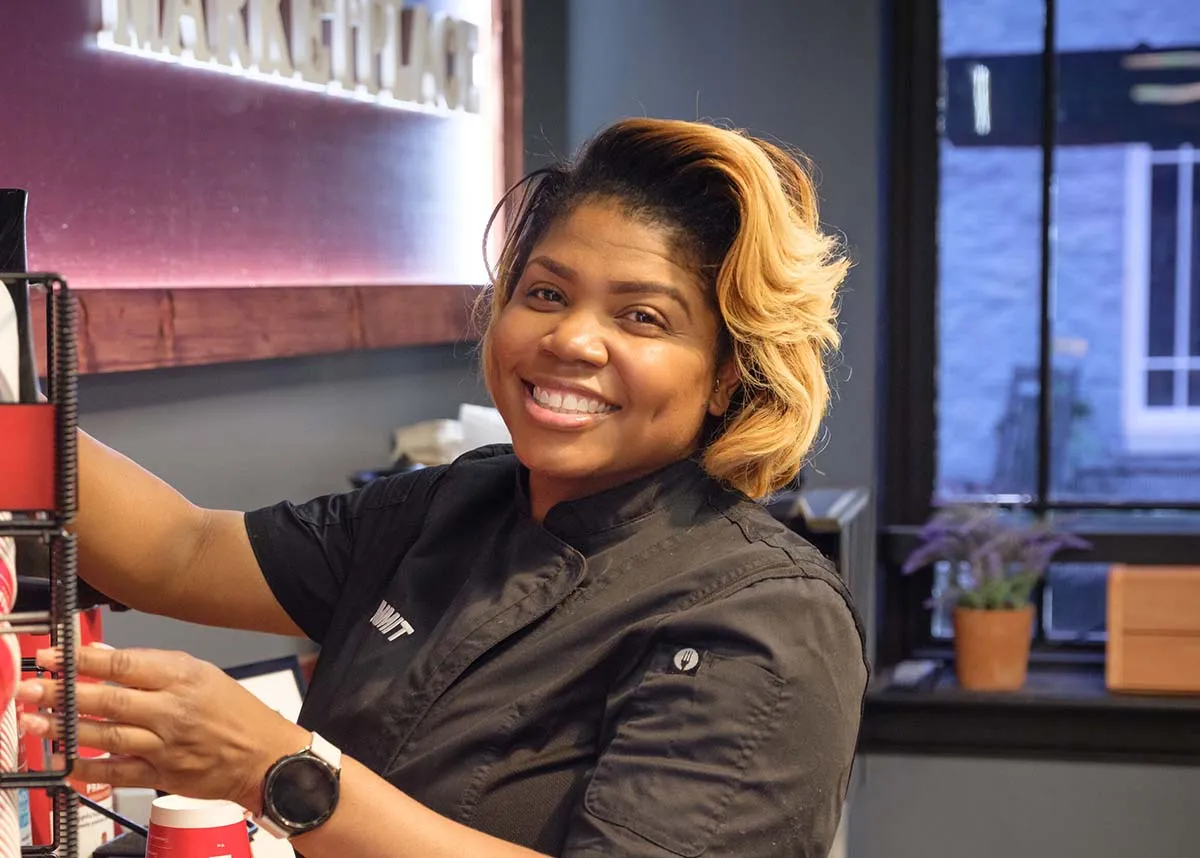 A smiling barista arranges coffee cups at a café counter with natural light from a window behind her.
