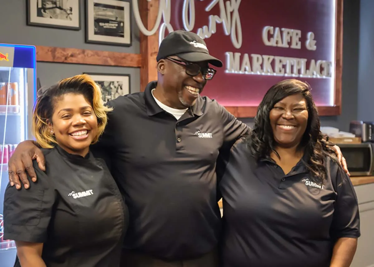 Three smiling people in matching black shirts pose together inside a cozy café and marketplace.