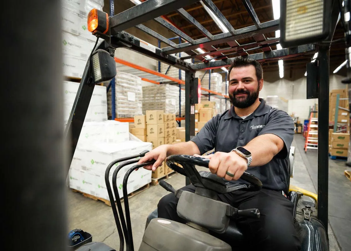 Smiling man operating a forklift inside a warehouse filled with stacked boxes and pallets.
