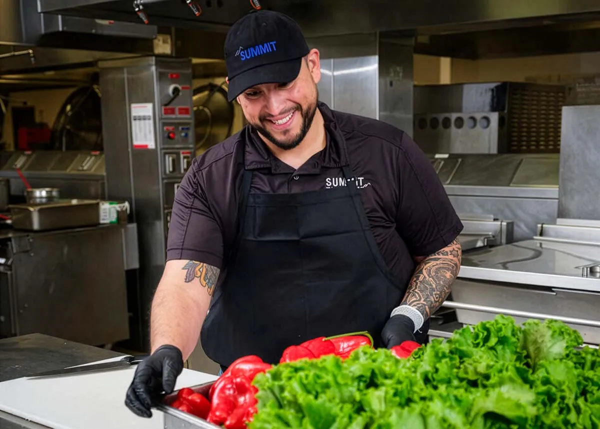 Smiling chef wearing a black apron and cap, handling fresh red bell peppers and leafy lettuce in a kitchen.