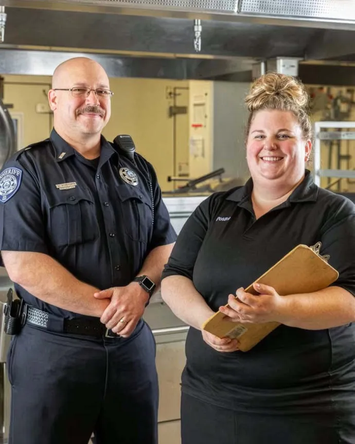 A police officer and a staff member standing and smiling together in a commercial kitchen.