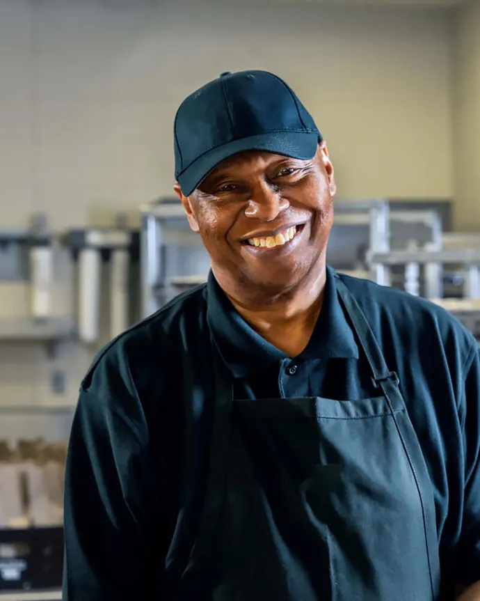 A smiling man wearing a black cap, black shirt, and black apron standing in a kitchen or food preparation area.