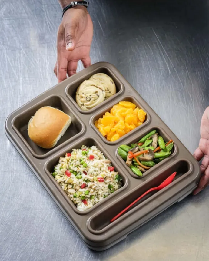 A person holding a compartmentalized tray containing a bread roll, a cinnamon roll, mandarin orange slices, a mixed vegetable stir-fry, and a grain salad, with a red plastic fork placed in an empty section.