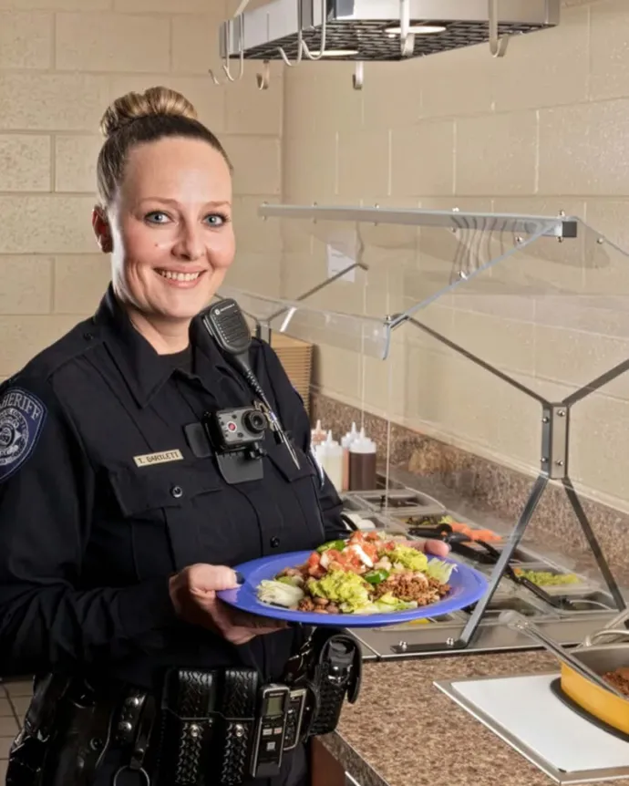 A smiling female police officer holding a plate of food in what appears to be a cafeteria or food service area.