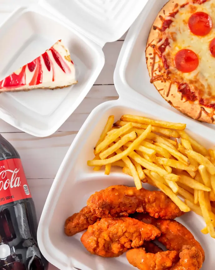 A meal consisting of fried chicken tenders, French fries, a pepperoni pizza, a slice of strawberry cheesecake, and a bottle of Coca-Cola, all set on a light wooden surface.