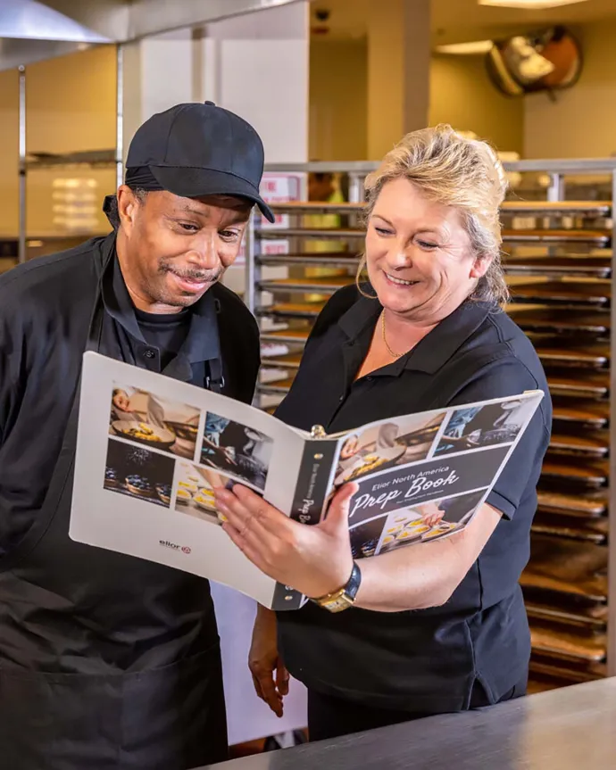 A man and a woman, both dressed in black work attire, are standing together in a kitchen and looking at a recipe book while smiling.